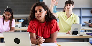 Schülerin sitzt an einem Tisch, vor ihr ein Laptop und Schreibutensilien. Im Hintergrund sitzen drei weitere Personen, von denen sich eine meldet.