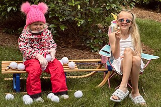 Ein Mädchen sitzt in Winterkleidung, mit Mütze und Schneehose, auf einem Schlitten. Neben ihr sitzt ein zweites Mädchen in sommerlicher Kleidung und mit Sonnenbrille und isst ein Eis.