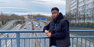 Junger Mann steht auf einer Autobahnbrücke. Im Hintergrund kann man ein blaues Autobahnschild erkennen.