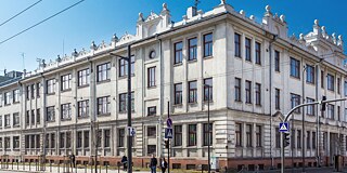 Gebäude von außen an einer befahrenen Straße, zwei Fußgänger stehen vor der Schule an der Ampel