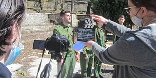 Schülerin hält Filmklappe vor Videokamera, drei Schüler in Uniformen stehen in einer Reihe