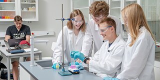 Vier Schüler und Schülerinnen in weißen Kitteln führen ein Chemieexperiment durch