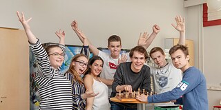 Schüler und Schülerinnen stehen um einen Hochtisch mit einem Schachspiel herum und lachen in die Kamera