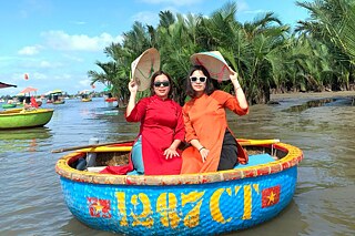 Zwei Frauen, in einem roten und einem orangenen Gewand, sitzen in einem runden, blauen Boot. Sie halten runde, spitz zulaufende Hüte über ihre Köpfe. Im Hintergrund sind Palmen zu sehen.