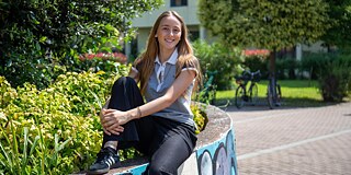 Eine junge Frau mit langen, blonden Haaren sitzt auf einer kleiner Mauer in einem Garten.