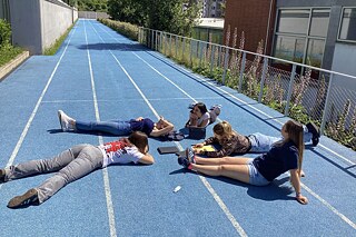 Mehrere Schülerinnen liegen auf einem asphaltierten Boden auf dem Bauch zueinander gewandt. Die Sonne scheint.