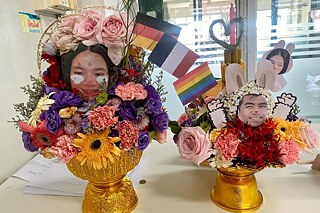 Zwei goldene Vasen mit bunten Blumen stehen auf einem Schreibtisch. In die Blumen sind zwei Fotos gesteckt, von einem Mann und einer Frau.