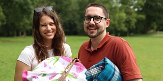 Zwei junge Erwachsene stehen auf einer grünen Wiese und halten eine Picknickdecke, einen Picknikkorb und eine Tischdecke in der Hand.