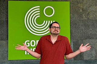 Ein junger Mann mit Bart und Brille steht vor einem Logo des Goethe-Instituts.