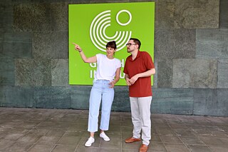 Zwei Erwachsene steht vor einem Logo des Goethe-Instituts, die Frau zeigt mit dem Finger nach links.