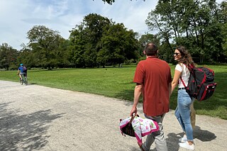 Zwei junge Erwachsene auf einem befestigten Weg in einem Park. Sie trägt einen offenbar schweren, schwarz7roten Rucksack, er einen leichten Korb, dessen Inhalt mit einer Decke zugedeckt ist.