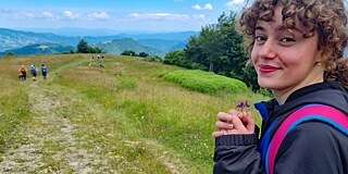 Eine junge Frau auf einem Wanderweg hält eine kleine Blume in der Hand und lächelt. Im Hintergrund sind Hügel und Bäume zu sehen.