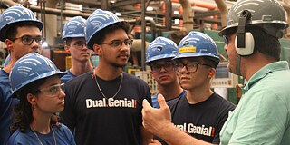 Sechs Jugendliche mit blauen Schutzhelmen und T-Shirts mit der Aufschrift „Dual-Genial“ hören einem Erwachsenen interessiert zu.