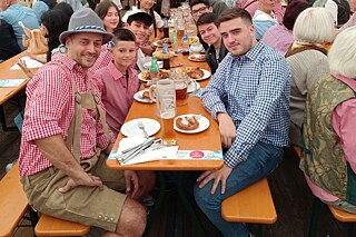 Zu sehen sind mehrere Personen, die in bayrischer Tracht an einer vollbesetzten Bierbank sitzen und Bierkrüge und Essen vor sich stehen haben. Sie sitzen in einem vollen Zelt, das mit blau-weißer Deko geschmückt ist.