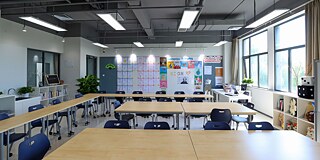 Leeres Klassenzimmer mit hellen Tischen und dunkelblauen Stühlen. An der Seite weiße Regale mit Büchern, Pflanzen und großen Würfeln aus Stoff. An der Wand sind geschriebene Seiten angebracht sowie verschiedene Poster und Bilder zum Thema "Economics"