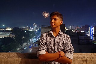 Eine Person posiert mit verschränkten Armen vor einer Mauer. Im Hintergrund ist ein Feuerwerk über einer Stadt zu sehen. Es ist dunkel und die Gebäude sind hell erleuchtet.