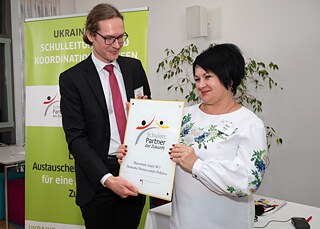 Ein Mann im schwarzen Anzug und eine Frau in einem weißen Oberteil stehen vor einem grünen Goethe-Institut-Aufsteller und halten gemeinsam eine Plakette, die die Aufschrift "Schulen: Partner der Zukunft" trägt.