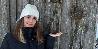 Eine junge Frau warm gekleidet mit heller Strickmütze steht vor einer Holzwand an der Hufeisen mit der Öffnung nach oben genagelt sind.