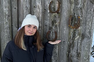 Eine junge Frau warm gekleidet mit heller Strickmütze steht vor einer Holzwand an der Hufeisen mit der Öffnung nach oben genagelt sind.