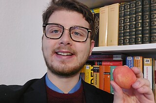 Ein junger Mann mit Brille steht vor einem Bücherregal; er zeigt ein rot gefärbtes Ei in die Kamera.