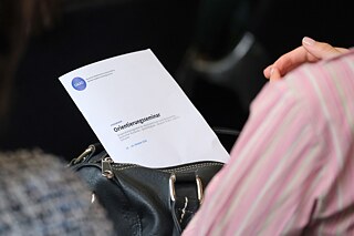 Eine Person in einer hellrosanen Bluse hat eine schwarze Handtasche vor sich. Diese ist geöffnet und ein Flyer mit dem Titel Orientierungsseminar ragt heraus.