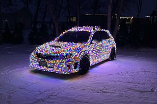 Ein Auto ist geschmückt mit vielen bunten Lichterketten. Sie bedecken das gesamte Auto. Im Hintergrund sind kahle Bäume zu erkennen. Es liegt Schnee.