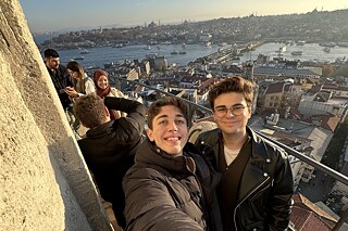 Zwei Personen stehen für ein Selfie zusammen auf einer Aussichtsplattform auf einem Turm und lächeln nach oben in die Kamera. Im Hintergrund sind Gebäude und ein Fluss zu sehen.