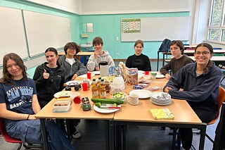 Sieben Schülerinnen und Schüler sitzen an einem großen Holztisch, auf dem gestapelte Teller, Geschirr, Getränke und Essen stehen.