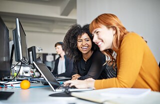 Zwei junge Frauen sitzen am Schreibtisch und arbeiten am Computer und Laptop. Sie unterhalten sich und lachen.  Im Hintergrund arbeitet ein Mann konzentriert, er trägt einen Kopfhörer. Er ist unscharf abgebildet.