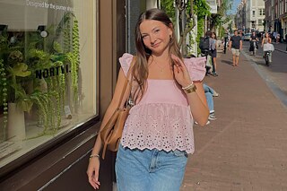 Eine junge Frau auf einer Einkaufsstraße, sie trägt eine rosa Bluse mit Volants und einen Jeansrock.