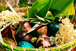 Foto eines geflochtenen Korbs mit grüner Bordüre. Im Korb befindet sich an den Seiten ein dekoratives grünes Blatt und eine gelbe Blume. Dazwischen liegen mehrere bunt gefärbte Eier.